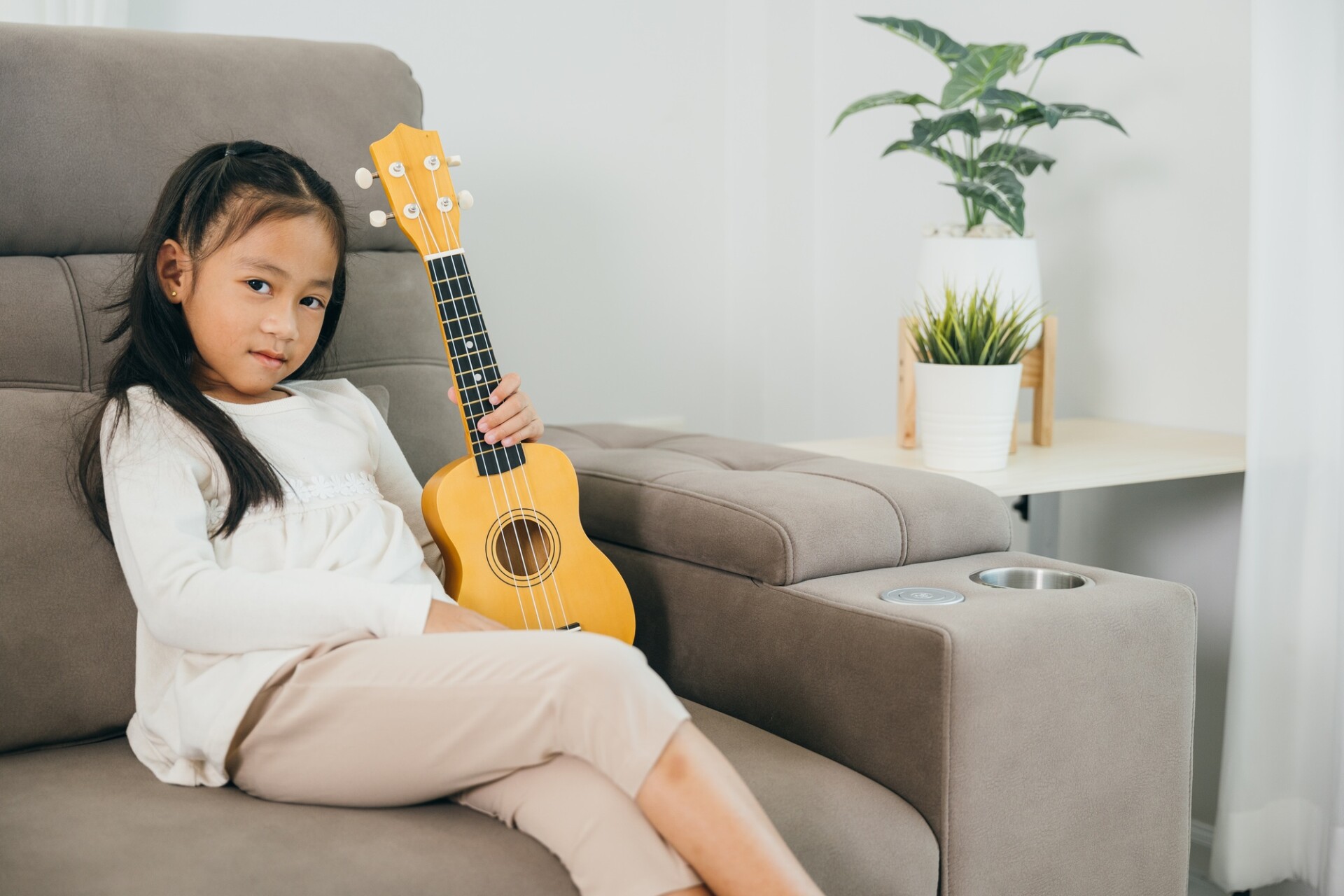 A young girl in a white shirt sits on a grey couch, holding a small wooden ukulele. Her bright smile